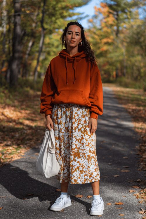 a woman dressed in a cozy burnt orange pullover hoodie with raw hem detailing, relaxed flowy floral printed midi skirt in warm autumn tones, no-show socks and white leather sneakers