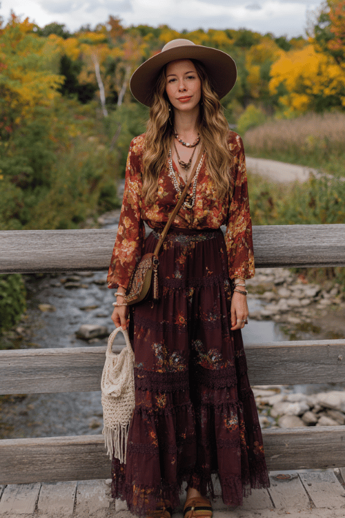 a woman dressed in a sheer floral printed boho blouse in warm autumn hues tucked into a long tiered skirt in deep plum with lace hem detailing, a floppy suede hat, layered pearl and crystal necklace