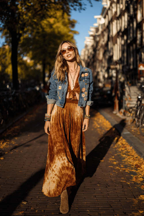 a woman in a vintage-inspired hippie chic outfit featuring a long tie-dye maxi dress in amber and brown tones, a cropped denim jacket with floral patches