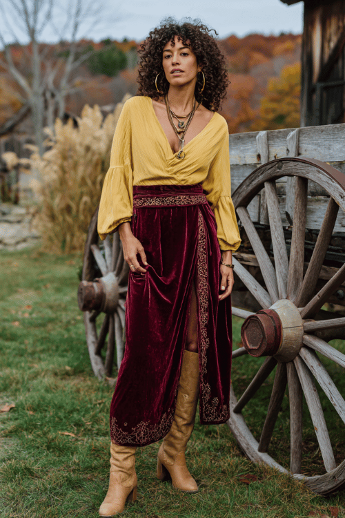 a woman wearing a rich burgundy velvet maxi skirt with a gold embroidered hem, a mustard yellow wrap top with deep V-neck and billowing sleeves