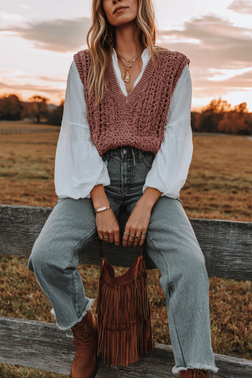 a woman wearing a soft dusty rose crochet vest over a long-sleeve white boho blouse with billowing sleeves, tucked into straight-leg medium wash jeans, lace-up flat leather boots