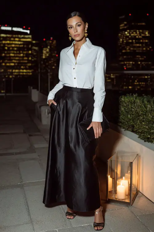 an elegant woman dressed in a classic crisp white tuxedo shirt with French cuffs and gold cufflinks tucked into a high-waisted floor-length satin skirt in deep midnight black