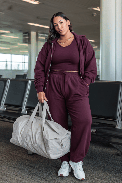 plus-size woman wearing a matching jogger set in deep burgundy a relaxed zip-up sweatshirt and wide-leg jogger pants with cushioned white sneakers and a large puffer tote bag 