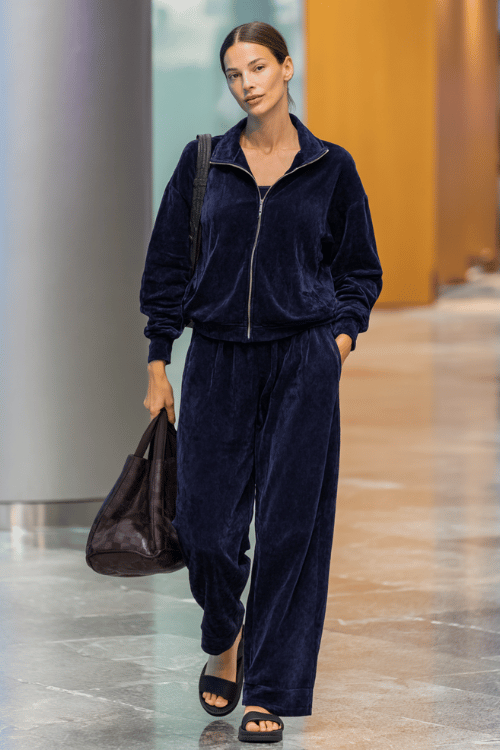 woman dressed in a velvety tracksuit in deep navy a relaxed zip-up top and wide-leg pants with thick cushioned slide sandals and a roomy leather tote bag