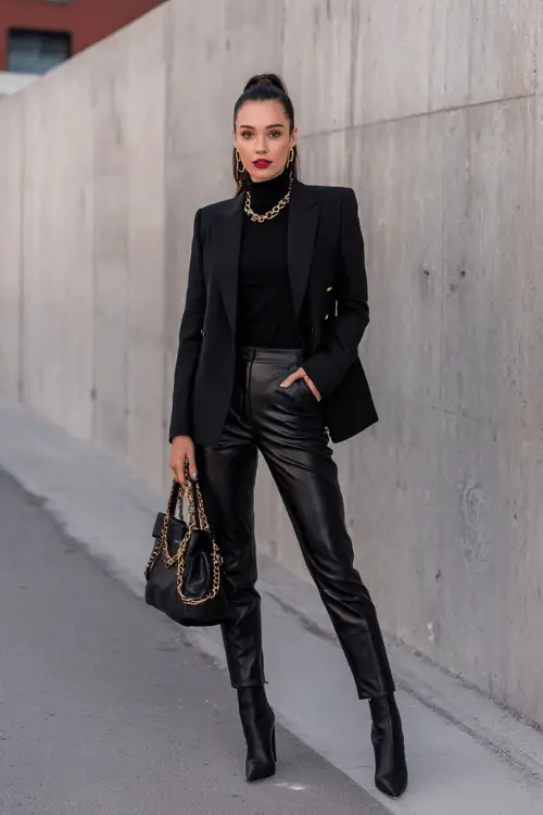 woman in her 40s wearing a classic tailored all-black ensemble featuring a blazer with gold chain lapel detailing over a fitted black turtleneck and straight-leg leather trousers 