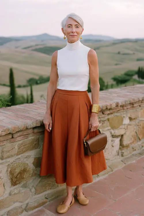 woman in her 50s wearing a classic sleeveless turtleneck knit top in crisp white tucked into a high-waisted A-line midi skirt in soft terracotta linen