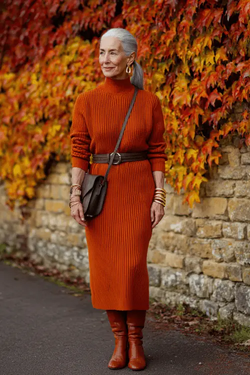 woman in her mid-60s dressed in a classic soft rust orange ribbed knit midi dress with long sleeves and a relaxed turtleneck neckline, layered with a tailored dark brown leather belt at the waist