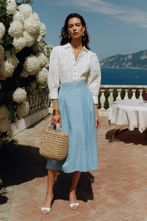 woman wearing a classic white broderie anglaise long-sleeve blouse with delicate eyelet detailing tucked into a high-waisted A-line midi skirt in soft sky blue linen