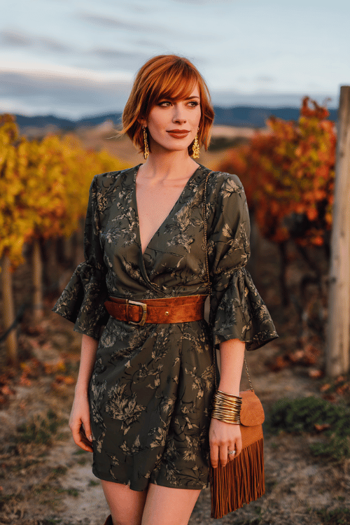 woman with a chic auburn bob wearing a deep olive wrap dress with botanical print and flutter sleeves, a structured leather belt, stacked gold bangles and drop earrings