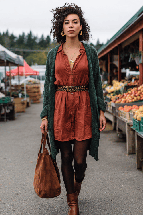 woman with natural dark curly hair wearing a relaxed rust-colored linen shirt dress belted at the waist with a braided leather belt, opaque dark tights, a long open-front boho cardigan