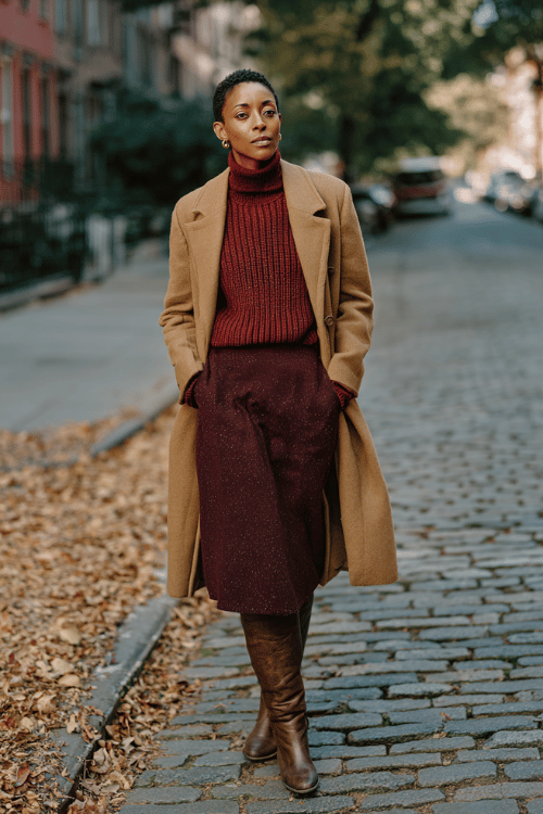 a Black woman dressed in a rich burgundy knit midi skirt and a matching ribbed turtleneck, layered with a long camel overcoat, wearing knee-high leather boots