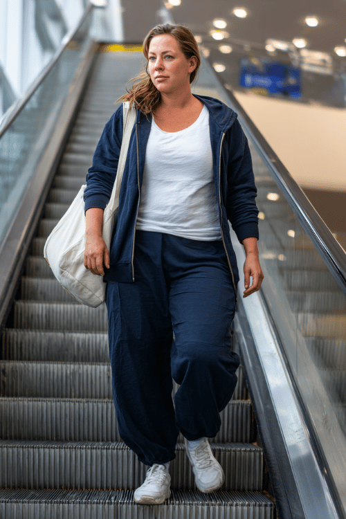 a plus-size woman ascending an airport escalator, wearing a soft navy blue zip-up track jacket, matching wide-leg track pants, a white fitted tee underneath 