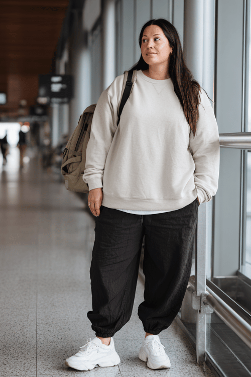 a plus-size woman at a modern airport terminal, wearing a soft oatmeal oversized crewneck sweatshirt, wide-leg dark jogger pants, and fresh white chunky sneakers