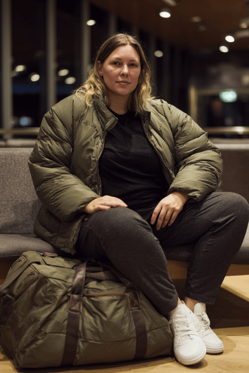 a plus-size woman at an airport lounge, wearing a forest green puffer jacket, black mock-neck top, slim-fit charcoal joggers, and white sneakers, duffle bag on her lap, cozy and casual chic