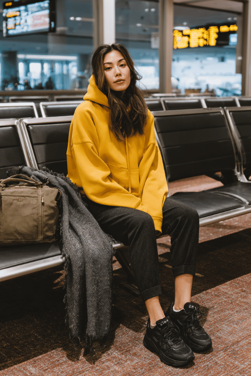 a woman at an airport gate seating area, dressed in a mustard yellow oversized hoodie, dark slim jogger pants, and black chunky sneakers, large cozy blanket scarf draped over her carry-on