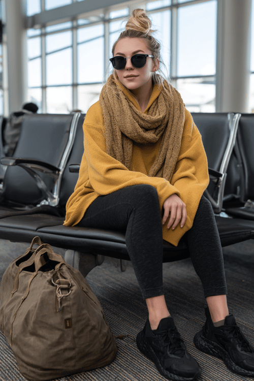 a woman at an airport gate seating area, dressed in a mustard yellow oversized hoodie, dark slim jogger pants, and black chunky sneakers