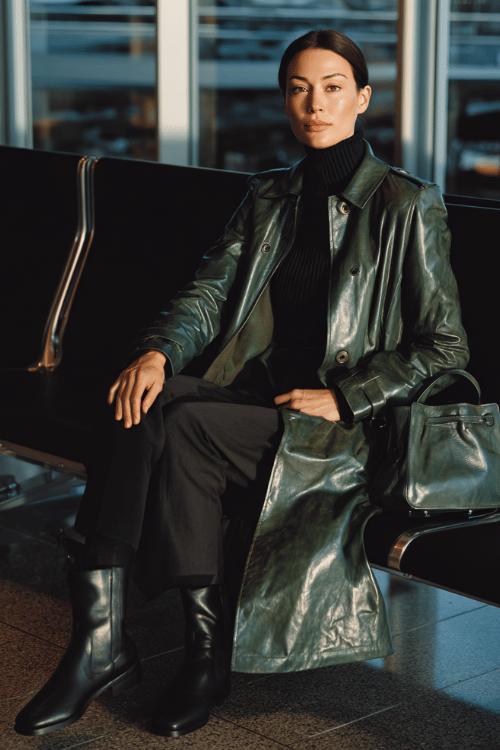 a woman at an airport lounge, wearing a deep forest green leather trench coat, black ribbed turtleneck, slim-fit dark trousers, and black Chelsea boots, structured mini handbag on her lap