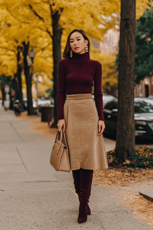 a woman in a deep burgundy ribbed turtleneck sweater tucked into a high-waisted A-line midi skirt in warm camel, wearing over-the-knee boots in dark brown suede 