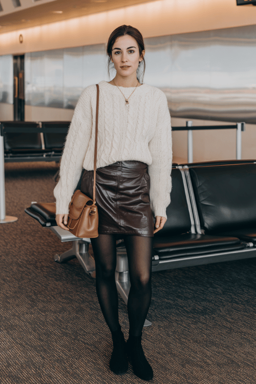 a woman waiting at an airport boarding gate, dressed in a rich chocolate brown faux leather mini skirt, ivory oversized knit sweater, opaque black tights, and black pointed-toe ankle boots 