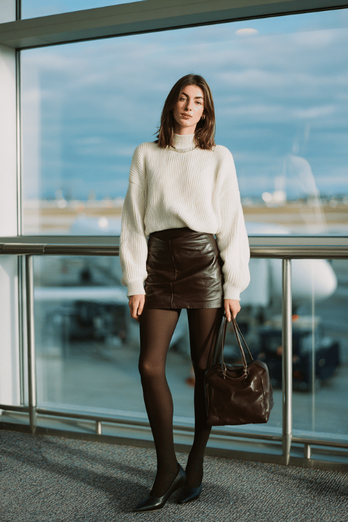a woman waiting at an airport boarding gate, dressed in a rich chocolate brown faux leather mini skirt, ivory oversized knit sweater, opaque black tights, and black pointed-toe ankle boots