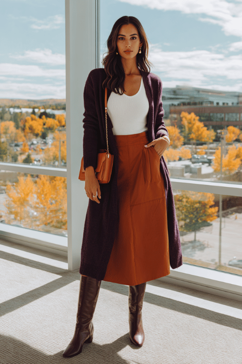 a woman wearing a deep plum longline cardigan over a white fitted camisole tucked into a high-waisted A-line midi skirt in warm rust-orange, finished with knee-high boots