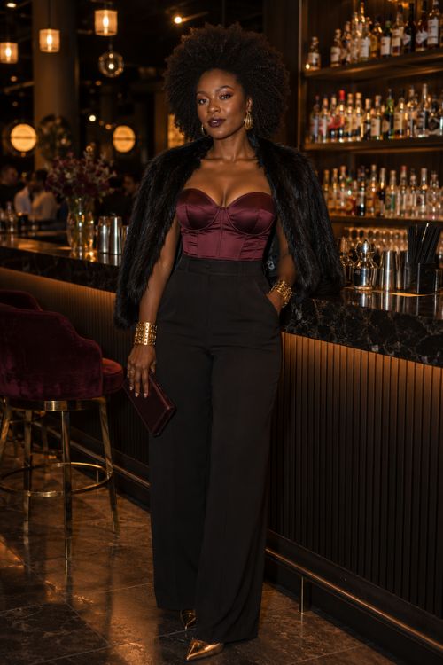 A Black woman wears a burgundy corset top, black wide-leg pants, faux-fur stole, gold heels, and a matching clutch