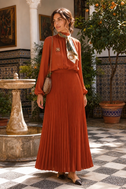 Woman in a rust pleated maxi skirt and scarf blouse in a Mediterranean courtyard.