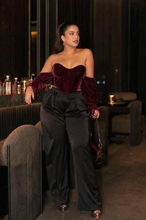 a woman wearing a burgundy velvet corset with a curved neckline, high-waisted black satin trousers, and metallic platform heels