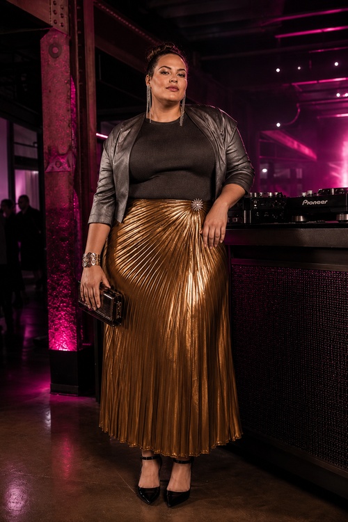 a woman wears a black top with a gold pleated midi skirt, a metallic jacket, and black pointed heels