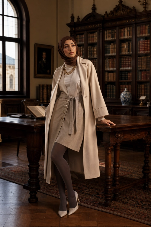 Elegant Beige Layered Outfit A woman wears a beige long coat over a light top and wrap skirt with dark tights, white pointed heels, a brown hijab, and a gold necklace.