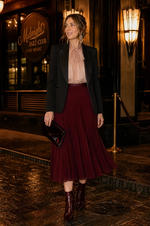 A tailored black blazer is styled with a blush pink ruffled blouse, burgundy pleated midi skirt, burgundy ankle boots, and a black clutch.