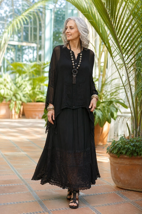 Black Bohemian Maxi Skirt Outfit A woman wears a black sheer long-sleeve blouse with a flowing lace-trimmed maxi skirt, layered necklace, bracelets, and flat sandals.