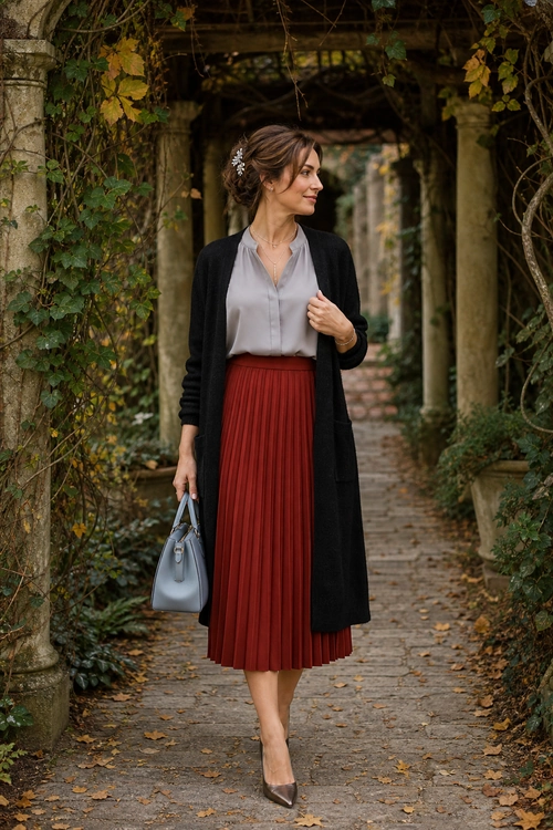 A woman wears a light gray blouse, long black cardigan, burgundy pleated midi skirt, and taupe pointed pumps while carrying a gray handbag.
