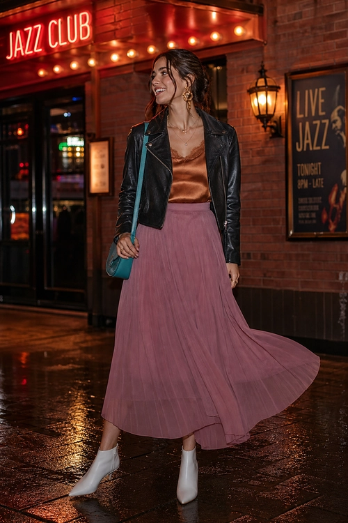 A woman wears a black leather jacket over a brown satin camisole with a flowing mauve skirt, teal shoulder bag, and white ankle boots.