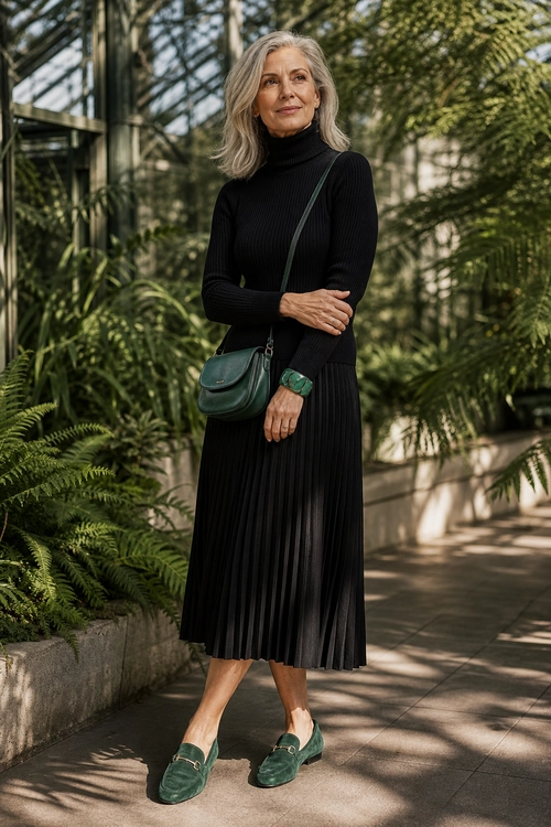 Black Pleated Dress with Green Accessories A woman wears a black ribbed turtleneck dress with a pleated midi skirt, green loafers, a green crossbody bag, and a green wristwatch.