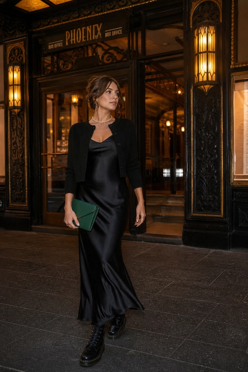 A woman wears a black satin maxi dress with a cropped black cardigan, black lace-up ankle boots, and a green clutch.