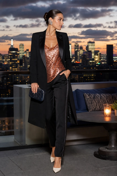 Black Tailored Evening Outfit A woman wears a long black blazer with black trousers, a copper sequin camisole, pointed cream heels, statement earrings, and a dark clutch.