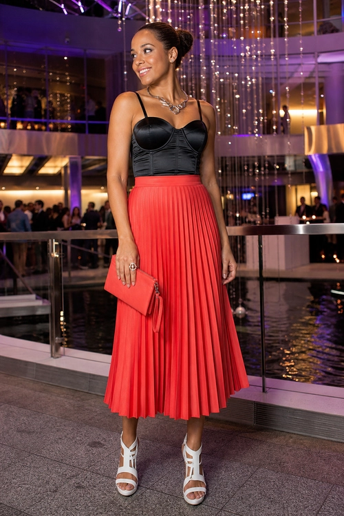 Woman wearing a black fitted top with a red pleated midi skirt, red clutch, and white strappy heels.