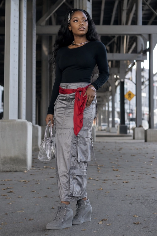 A woman wears a fitted black long-sleeve top with a silver cargo maxi skirt, red waist scarf, gray lace-up boots, and a clear handbag.