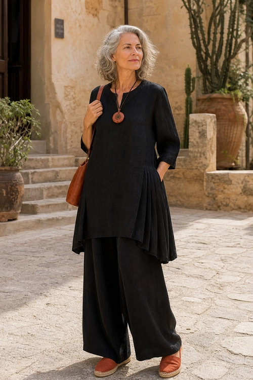 Black Tunic and Wide-Leg Trousers A woman wears a black long tunic with pleated side panels, wide-leg trousers, rust espadrille sandals, a brown shoulder bag, and a pendant necklace.
