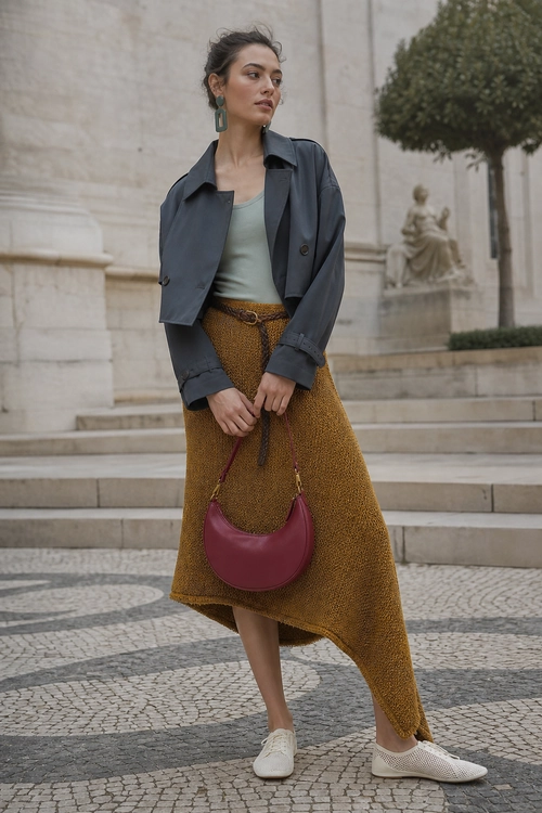 A woman wears a dark blazer, sage green top, asymmetric mustard skirt, burgundy shoulder bag, and white sneakers.