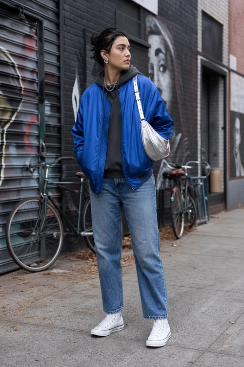 Street style outfit featuring a bright blue bomber jacket, dark hoodie, relaxed blue jeans, white high-top sneakers, and a silver shoulder bag.