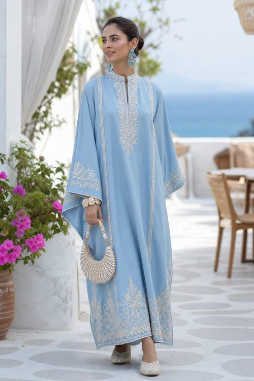 Woman wearing a flowing light blue embroidered kaftan with white flats, statement earrings, a beaded handbag, and bracelets.
