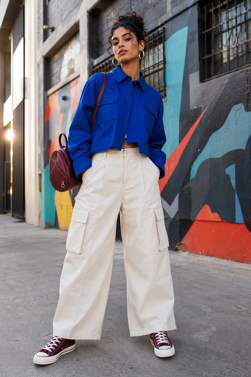 A person wears a cropped blue utility jacket with white wide-leg cargo pants, burgundy high-top sneakers, and a matching shoulder bag.