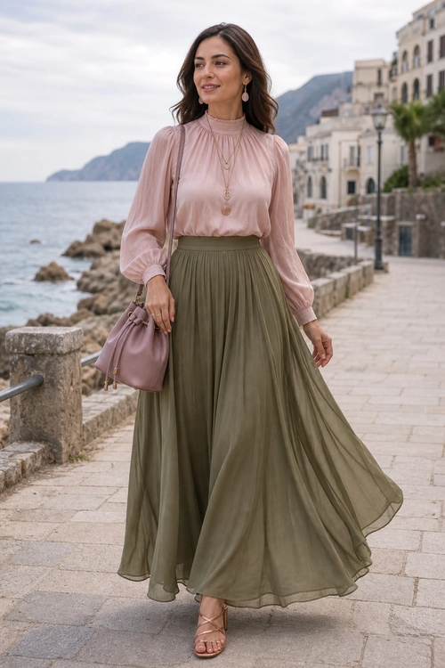 Blush Blouse and Olive Maxi Skirt Outfit Woman wearing a blush pink high-neck blouse with an olive green flowing maxi skirt, pink handbag, jewelry, and strappy sandals on a seaside promenade.