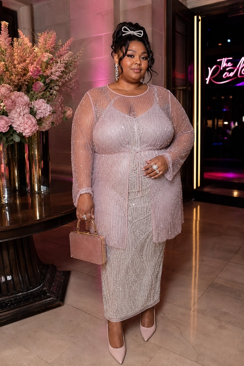 A woman wears a blush pink sparkly sheer long-sleeve overlay with a matching shimmering midi dress, pointed heels, and a structured clutch.
