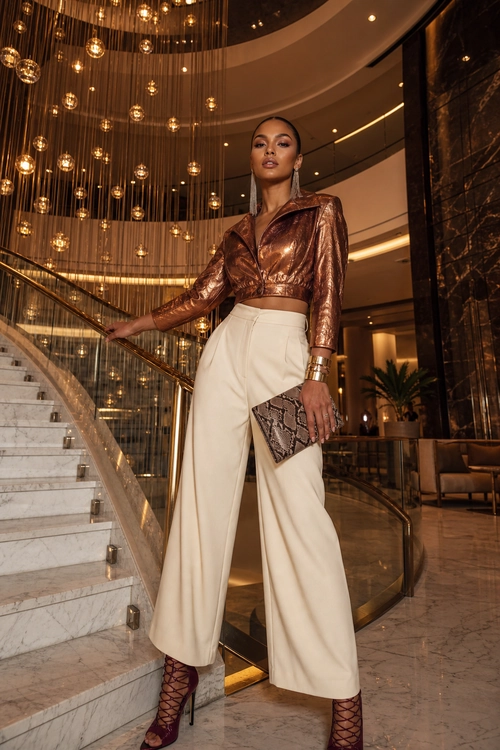 A woman wears a metallic bronze cropped long-sleeve jacket with ivory wide-leg trousers, burgundy lace-up heels, and a patterned clutch.