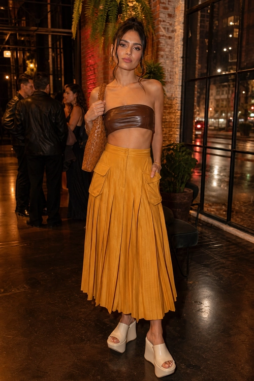 A woman wears a brown strapless bandeau top with a mustard pleated skirt, woven shoulder bag, and white platform sandals.