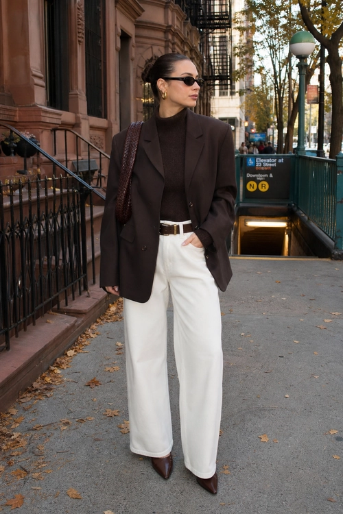 A woman wears a dark brown blazer over a brown high-neck top with white wide-leg trousers, pointed brown shoes, sunglasses, and a shoulder bag.