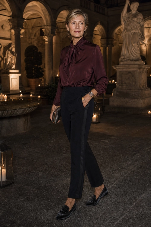 A woman wears a burgundy tie-neck blouse with black ankle-length trousers, black loafers, a slim belt, and a dark clutch.